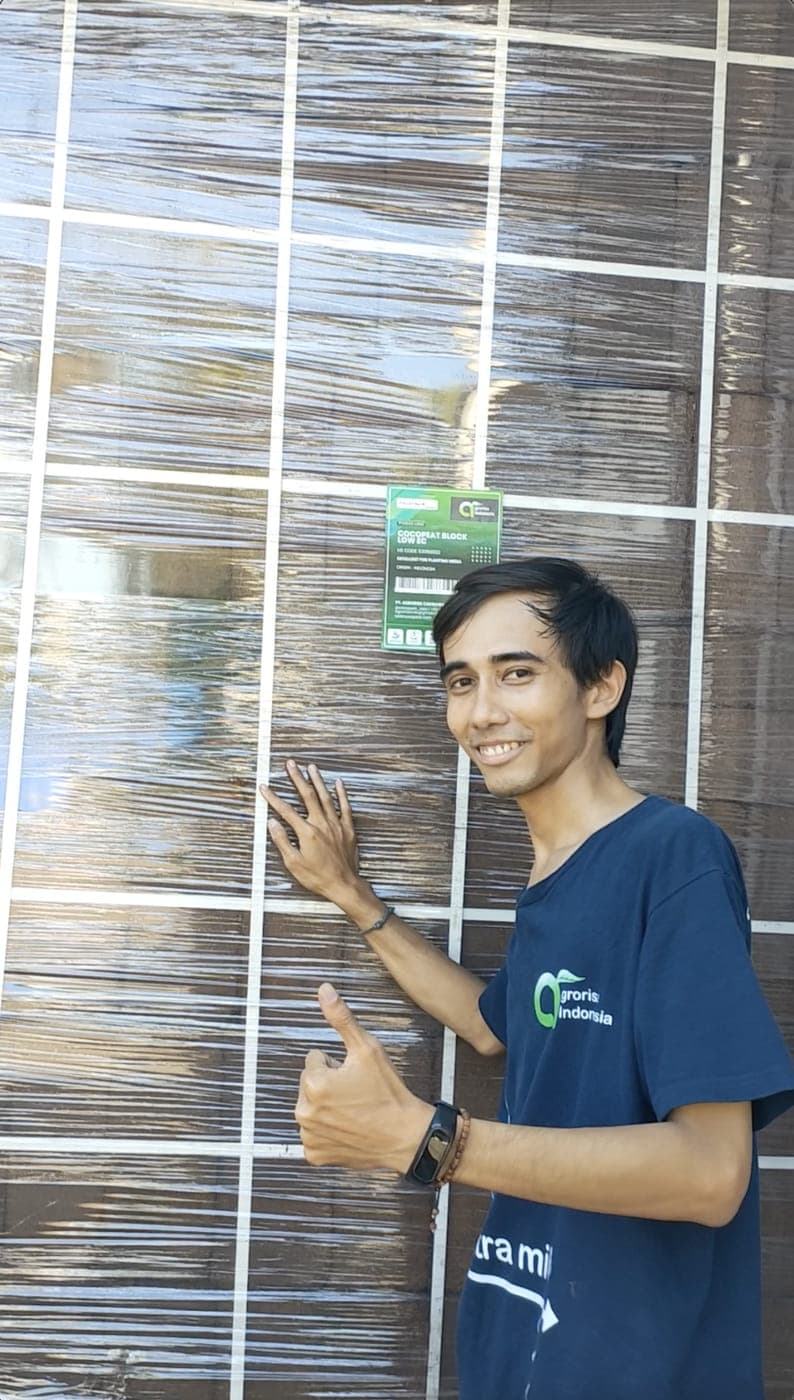 Agrorise Indonesia team member standing beside a tall wrapped pallet of cocopeat blocks ready for despatch.